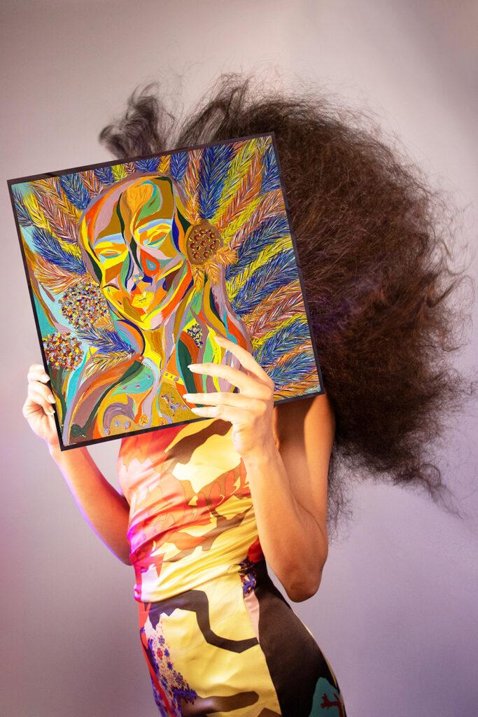La peintre Lila Ramos tient l'un de ses tableaux devant son visage comme si c'était son visage représenté en autoportrait.